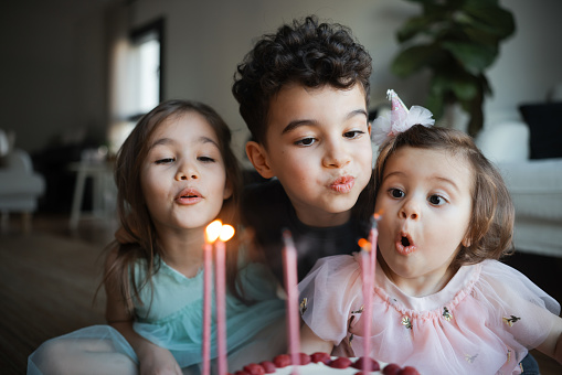 Child birthday party. Adorable little boy and girl with birthday cake decorated flowers and burn candle, makes wish. Kid in festive dress at room with balloons, bouquet of flowers at vase