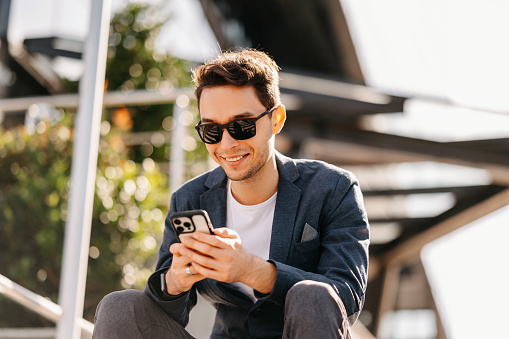 Young man wearing jacket and sunglasses is sitting outdoors using phone on a sunny day.