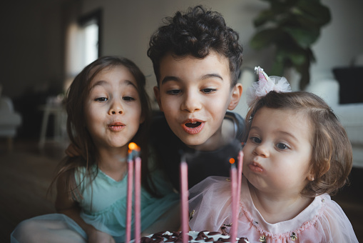 Child birthday party. Adorable little boy and girl with birthday cake decorated flowers and burn candle, makes wish. Kid in festive dress at room with balloons, bouquet of flowers at vase