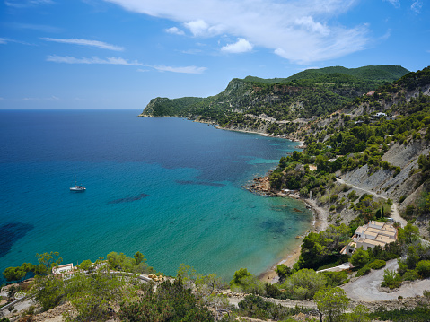 Wide-angle view of a long stretch of the southern coast of Ibiza, from the cove and beach of Platja des Cubells (at the bottom) to the verdant cliffs of Cap Llentrisca. The dazzling bright light of a Mediterranean summer noon, picturesque clouds, colourful cliffs covered with lush pine trees and a wide variety of Mediterranean shrubs and plants, scattered villas and holiday homes, a sailing boat rocking gently on crystal-clear waters. High level of detail, natural rendition, realistic feel. Developed from RAW.