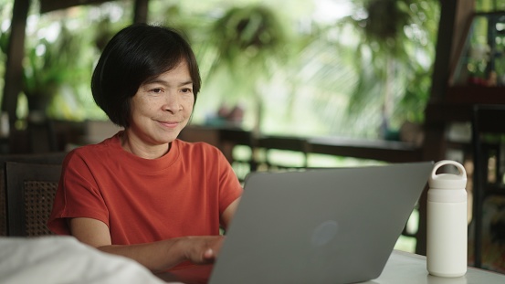 Asian mature woman working on laptop while resting at nature resort.