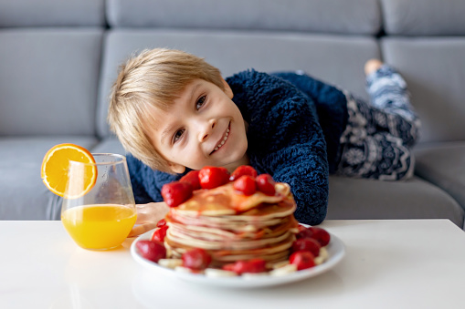 Sweet toddler child, boy, eating american pancakes with strawberries and bananas, topped with syrup and drinking fresh orange juice