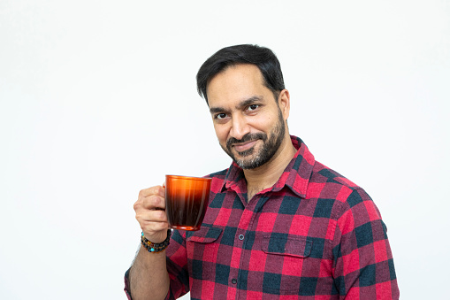 A cheerful man holding a red coloured coffee cup photographed against a plain background. He is wearing a red checked shirt.
