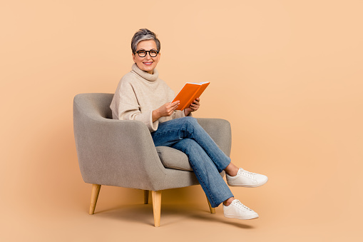Full length photo of happy old grandmother spending her pensioner like dream with book sitting armchair isolated on beige color background.