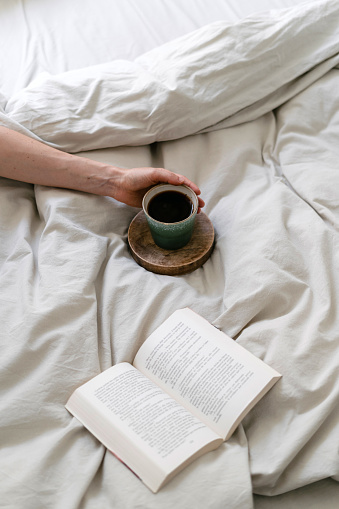 Cropped view on woman lying in comfort bed with rumpled blanket, hold cup of coffee with hand and reading open book in morning. Female resting at weekend in bedroom with soft bedding linen. Lazy mood