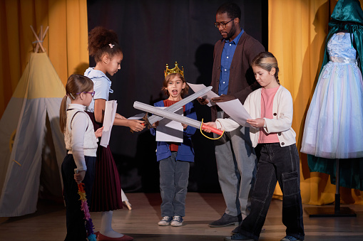 Full length portrait of children rehearsing school play on stage in theater and fighting with toy swords