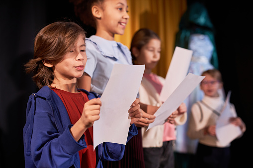 Side view portrait of young boy rehearsing lines standing on stage in school theater in row with children actors