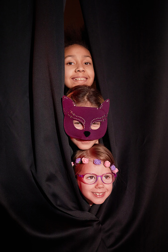 Three little child actors in costume peeking from curtain on stage in theater