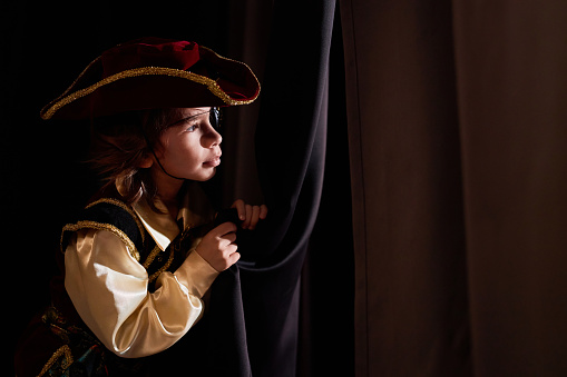 Side view portrait of little boy wearing costume peeking over curtain backstage in theater copy space