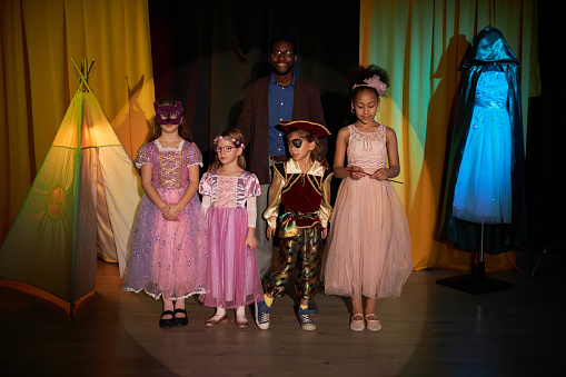 Full length portrait of group of little kids standing on stage in theater with smiling drama teacher