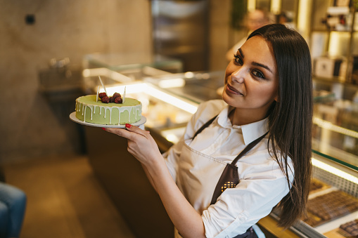 Beautiful young woman holding a cake and looking at camera while working in confectioner