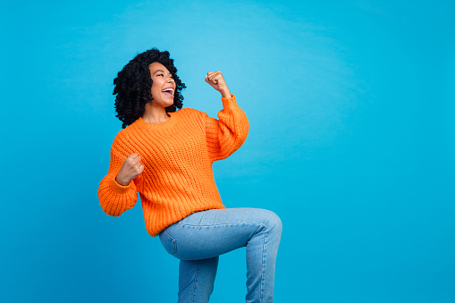 Photo of attractive young woman raise fists scream celebrate dressed stylish knitted orange clothes isolated on blue color background.