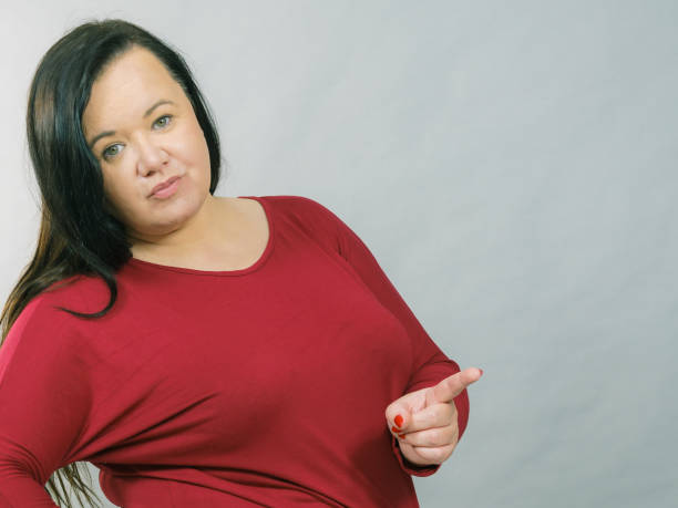 Adult plus size woman pointing with finger at copy space with funny face expression, on grey