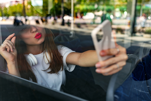 Woman with red lips sits on a city bus and takes a selfie using a smartphone. Photograph through a glass window.