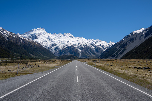 Discover the picturesque country road in New Zealand's South Island leading to the renowned Mount Cook National Park. Behold the stunning sight of snow-capped Mount Cook, majestically framed by other towering mountain peaks.