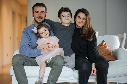 Happy young Caucasian family renters with two small kids sit rest on couch in cozy living room. Smiling parents with little children relax on sofa enjoy weekend in new renovated design home together.