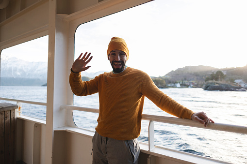 man leaning on the railing of a ferry on his vacation trip looking at camera and waving