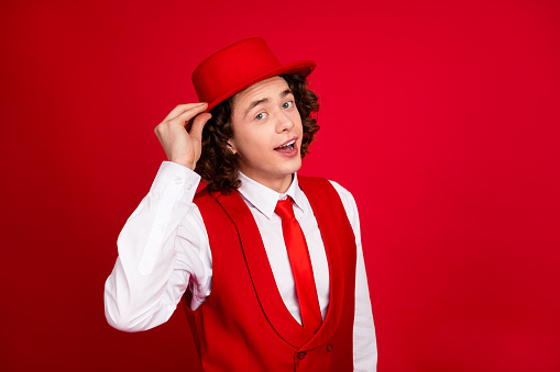 Portrait of handsome teenager guy wearing vest and shirt classy hat cone future showman posing model isolated on red color background.