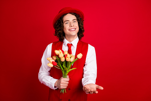 Photo of happy teenage boyfriend wearing formal suit gives you a hand invitation event and bunch tulips isolated on red color background.