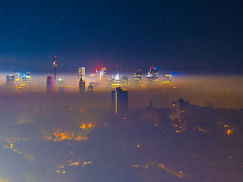 The photo depicts a scene of Sydney shrouded in heavy smog. The city lights are dimly visible through the dense fog, with the outlines of skyscrapers blurred and indistinct. Only the brightest lights manage to pierce the haze, adding a hint of vitality to the otherwise mysterious and hazy atmosphere. The scene evokes thoughts of unusual weather conditions or environmental pollution events.