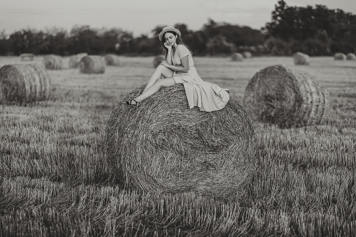 Portrait of a young girl. A girl in a blue dress sits on top of hay bales - high quality photo. Long straight hair. Nice color. Summer. Black and white photo