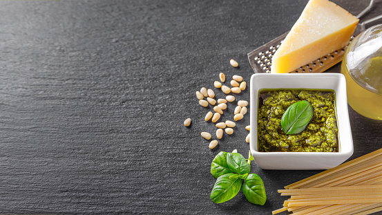 Homemade pesto sauce in small square jar and ingredients for pasta on black slate background with copy space. Traditional Italian cuisine, recipe, restaurant menu