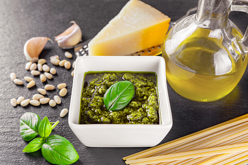 Homemade pesto sauce in small white square jar and ingredients for pasta on black stone background. Traditional Italian cuisine, recipe, restaurant menu