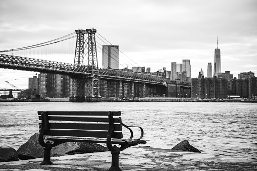 An empty rustic wooden bench with nostalgic Manhattan Skyline and Williamsburg Bridge on a snowy winter day, moody waterfront landscape over the East River and the Grand Ferry Park in Brooklyn, New york, USA, black and white photo