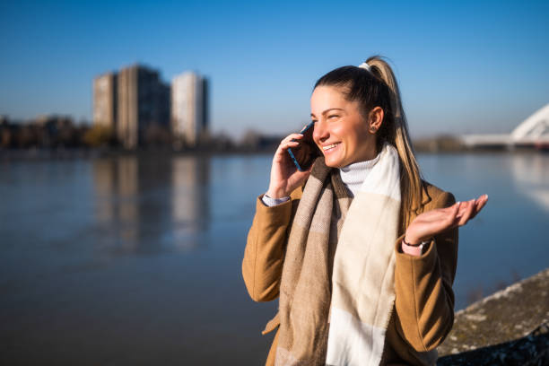 Beautiful woman in warm clothing talking on a mobile phone and enjoys resting by the river on a sunny winter day.