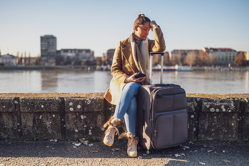 Worried woman tourist  with suitcase and phone lost in a city sitting by the river and thinking. Toned image.