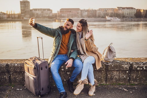 Happy couple tourists taking selfie while they are sitting and resting on  their vacation with their luggage.