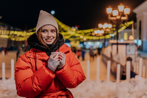 Cheerful young woman wearing warm winter clothes enjoying drinking takeaway cup of coffee outdoors on cold street at night with festive bright illumination. Pretty female walking on urban cold park