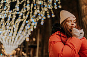 Portrait of pretty frozen young woman in hat and winter jacket warming hands in cold winter night standing posing on snow city street