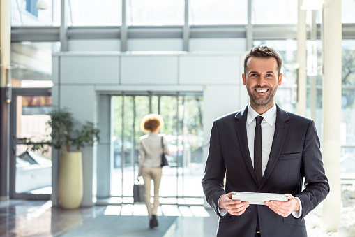 Portrait of happy businessman standing in hotel hall and holding a digital tablet in hands, smiling at camera.