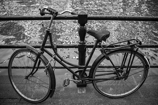 Black and white image of a bicycle parked and secured on the railing of a canal bridge in Amsterdam.