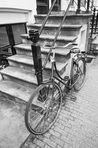 Black and white image of a bicycle parked at a fence at the steps of a traditional Amsterdam canal house.