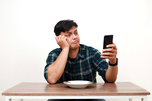 Confused young asian man choosing food on his phone while sitting with empty plate on the table. Online food order concept