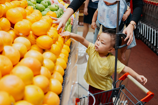 Little Asian Chinese boy taking fresh oranges from the produce aisle. Asian family grocery shopping for organic fruits and vegetables at supermarket.