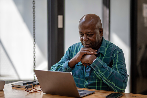 A black senior man in casual clothes is focusing on a laptop screen, concentrating and thinking seriously.