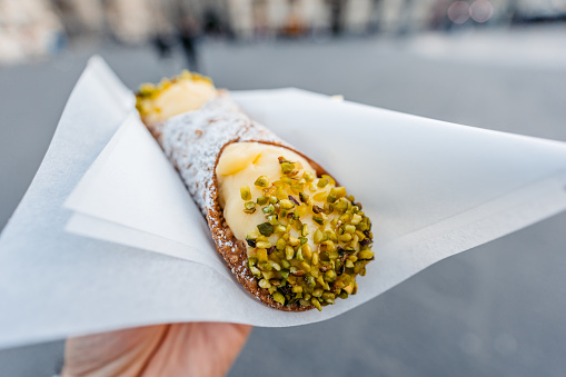 Young man holding a cannoli pastry filled with creamy ricotta and pistachios on the street in Catania, Sicily.