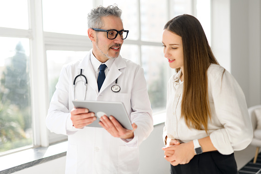 A grey-haired doctor in a white coat shares information on a tablet with a young woman in a bright, modern medical office, embodying a collaborative healthcare approach