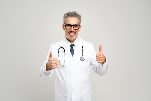 The senior doctor enthusiastic shows thumbs-up suggests a positive outcome or good news, complemented by his friendly expression and professional attire, against a grey backdrop.