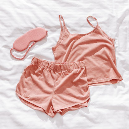 Top view pink pajama and eye sleep mask on white crumpled bedclothes. Cozy pyjamas for comfort rest at night. Flat lay from singlet, shorts, sleeping mask pastel pink color, sleep well concept