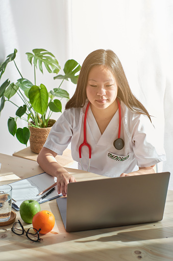 Focused woman in medical uniform sitting at wooden table and looking at laptop screen while working in modern clinic