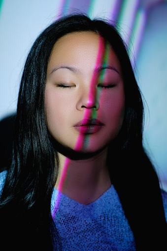Charming Asian female with long black hair standing near wall with closed eyes in room with colorful neon lights on face