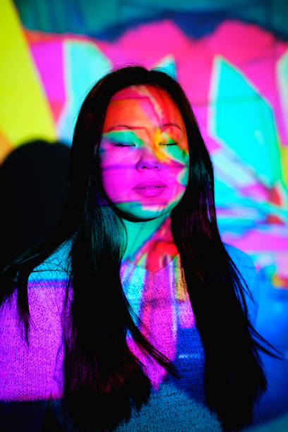 Tranquil Asian female with long black hair standing with closed eyes near wall under bright multicolored neon illumination in stylish room