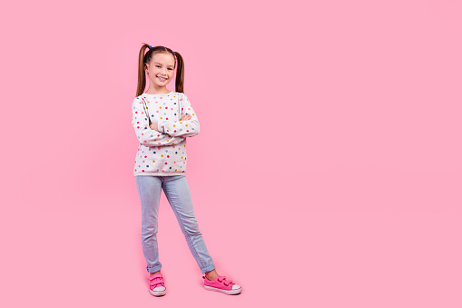 Full length photo of positive cheerful small girl wear stylish sweatshirt jeans hold hands crossed isolated on pink color background.