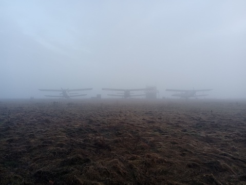 Three small sports planes are visible in the morning fog on the horizon. The planes are standing in an open field covered with morning fog.