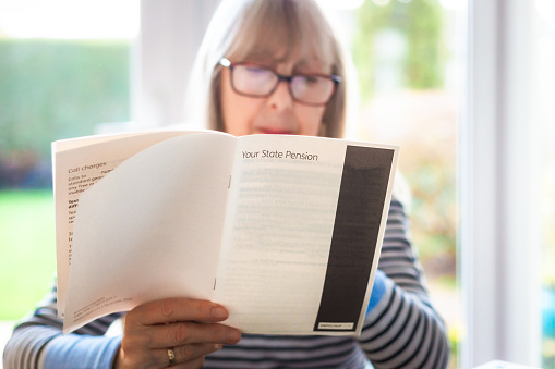 A retired senior woman in her 70s reads information about state pension. She is sitting in a room at home with the garden defocused outside. Focus on the leaflet with the woman slightly defocused in the background.
