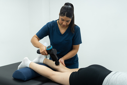 Providing restorative care, the young female physical therapist relieves tension in the patient's leg with an expert massage with muscle gun, promoting muscle relaxation and restoring balance for enhanced physical comfort.